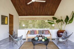 A modern living room with a wooden ceiling, a wall-mounted light fixture, a sofa with patterned cushions, and two wicker chairs. - caSabama (Blahbatuh)