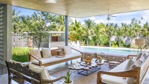 Amplia zona de estar al aire libre con piscina, mobiliario de madera y exuberante vegetación tropical.