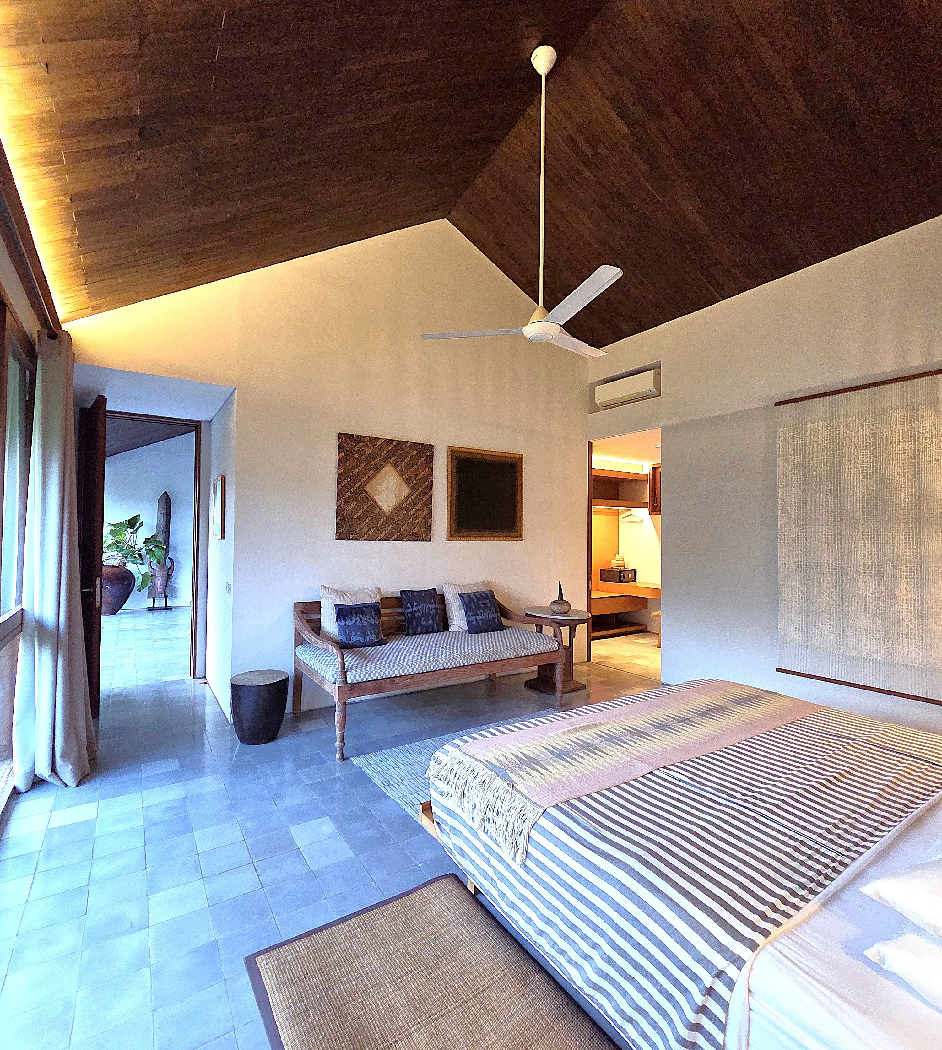 Une pièce spacieuse avec un grand lit, un banc en bois et un ventilateur de plafond. Une porte donne accès à une autre pièce, et une grande fenêtre laisse entrer la lumière naturelle.