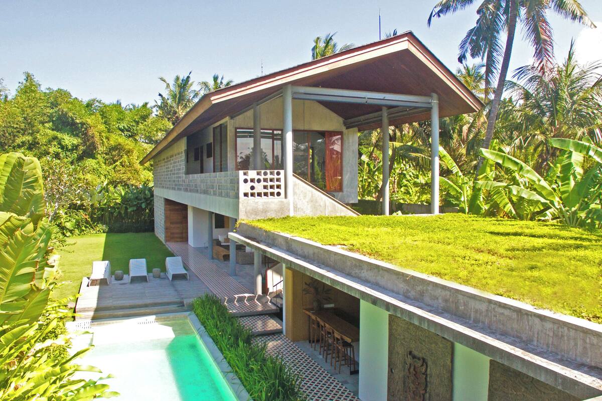 Una casa moderna con un porche cubierto, piscina y abundante vegetación.