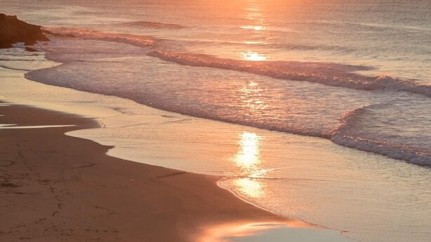 A beach scene with a sunset, waves, and two people walking with surfboards.