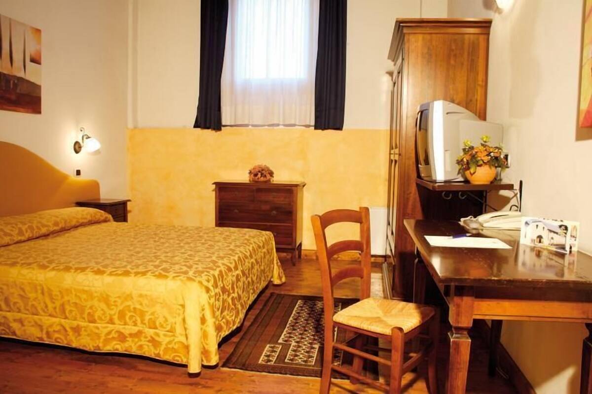 Hotelkamer met een bed, een bureau, een stoel, een klein kastje, een lamp en een raam met gordijnen.