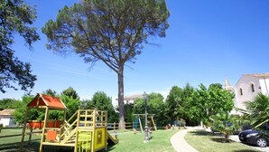 Un parque infantil con una estructura de juego de madera, un árbol grande y un sendero que lleva a un edificio con una torre de campanas.