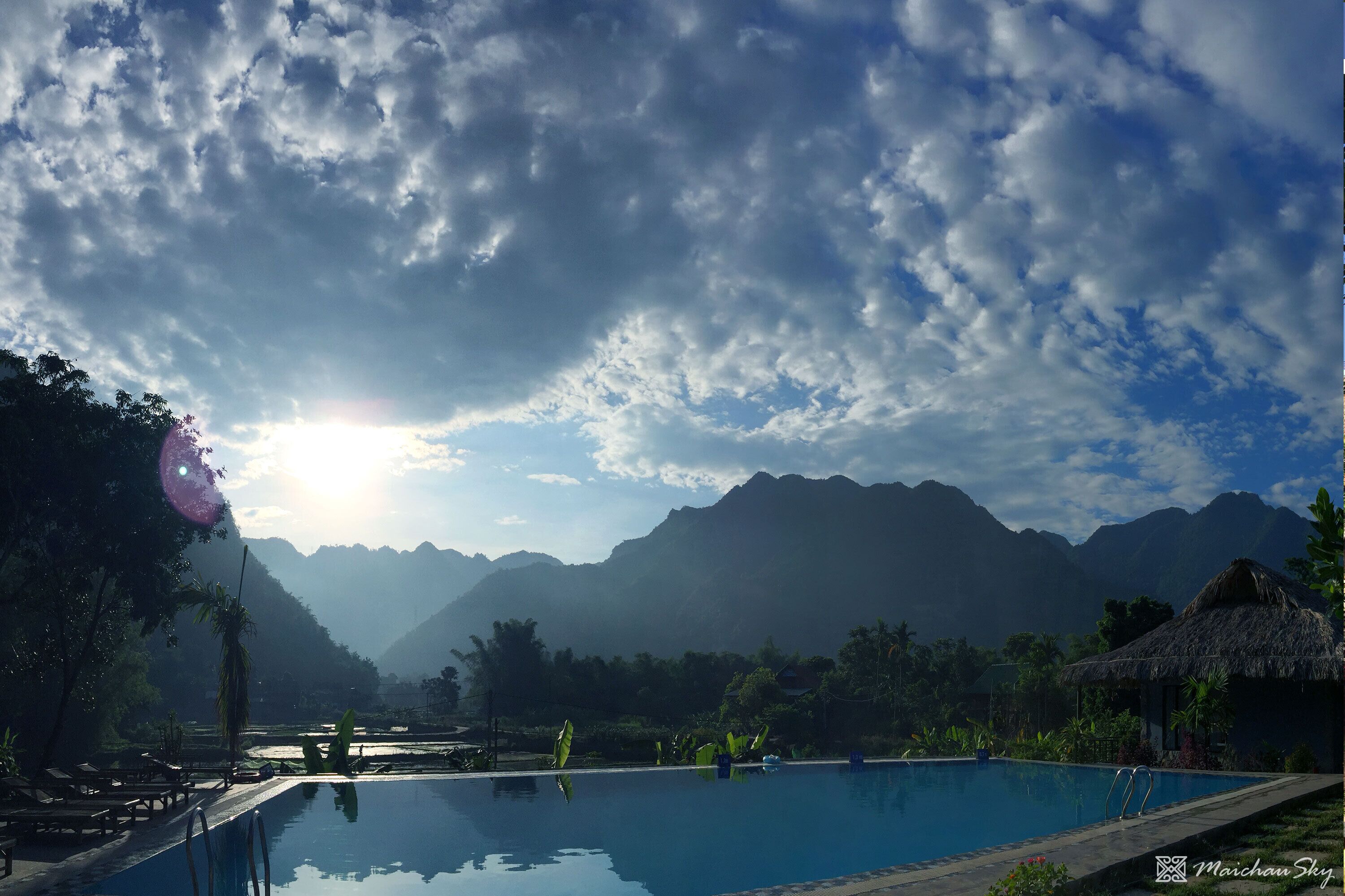 A pool with a mountain backdrop, surrounded by lush greenery and a thatched-roof structure.