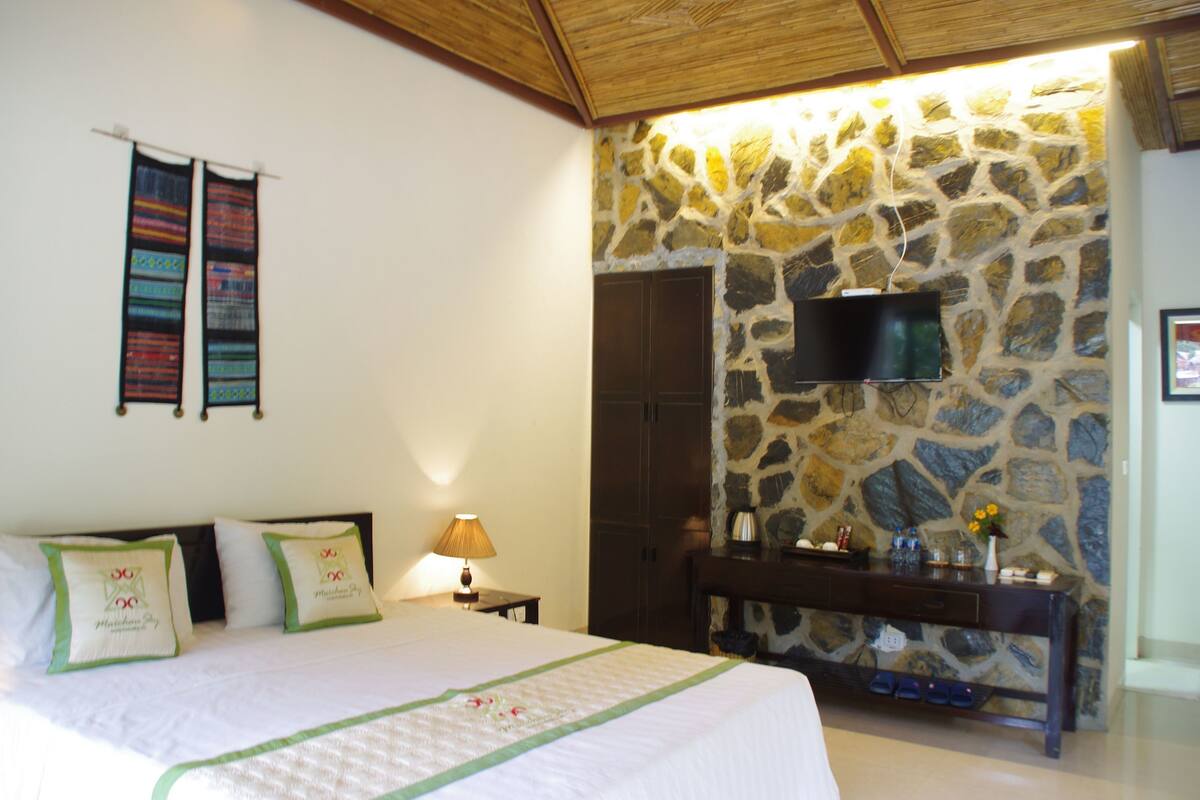 Une chambre avec un mur en pierre, un téléviseur à écran plat, un lit avec des oreillers verts et une table de chevet avec une lampe.