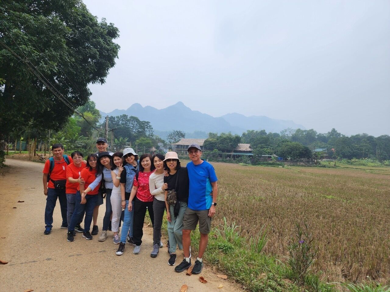 A group of people standing on a dirt path in a rural area with a field and mountains in the background.