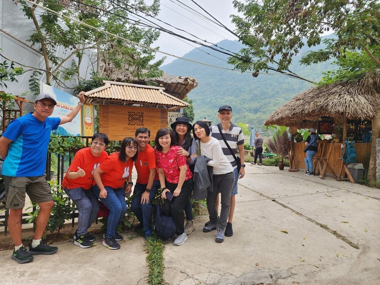 A group of people posing in front of a traditional wooden house with a thatched roof, surrounded by lush greenery and mountains in the background.
