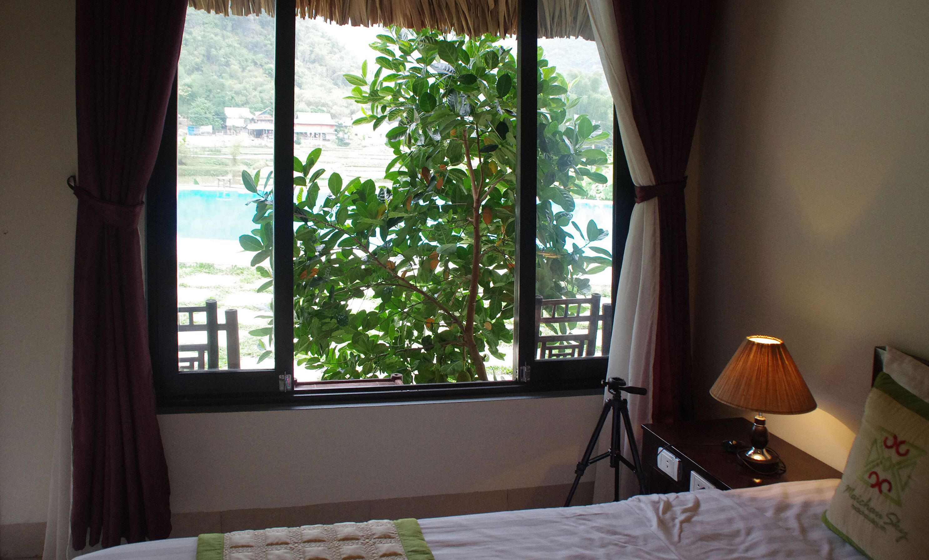 A hotel room with a bed, a desk with a lamp, a tripod, and a window with curtains.