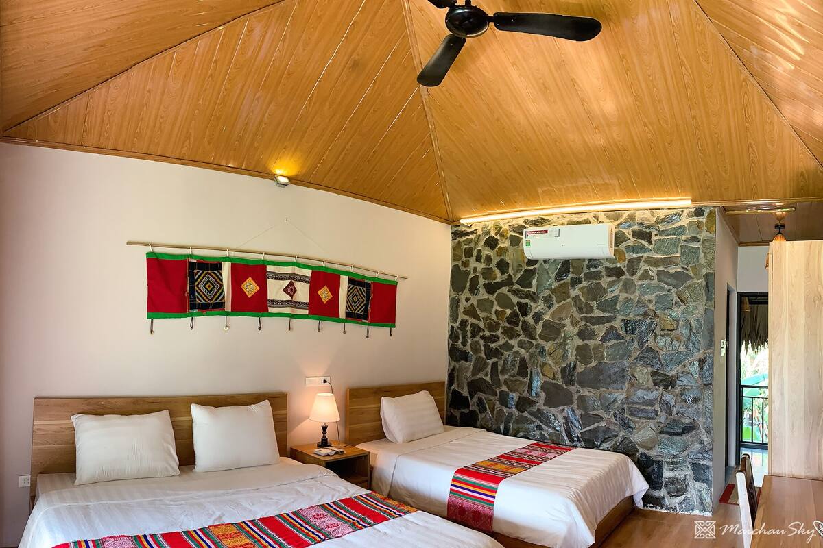 Une chambre avec deux lits, un mur en pierre, un ventilateur de plafond et un climatiseur fixé au mur.
