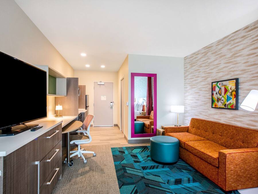 A modern hotel room with a flat-screen TV, a desk with a chair, a sofa, a blue ottoman, and a lamp.