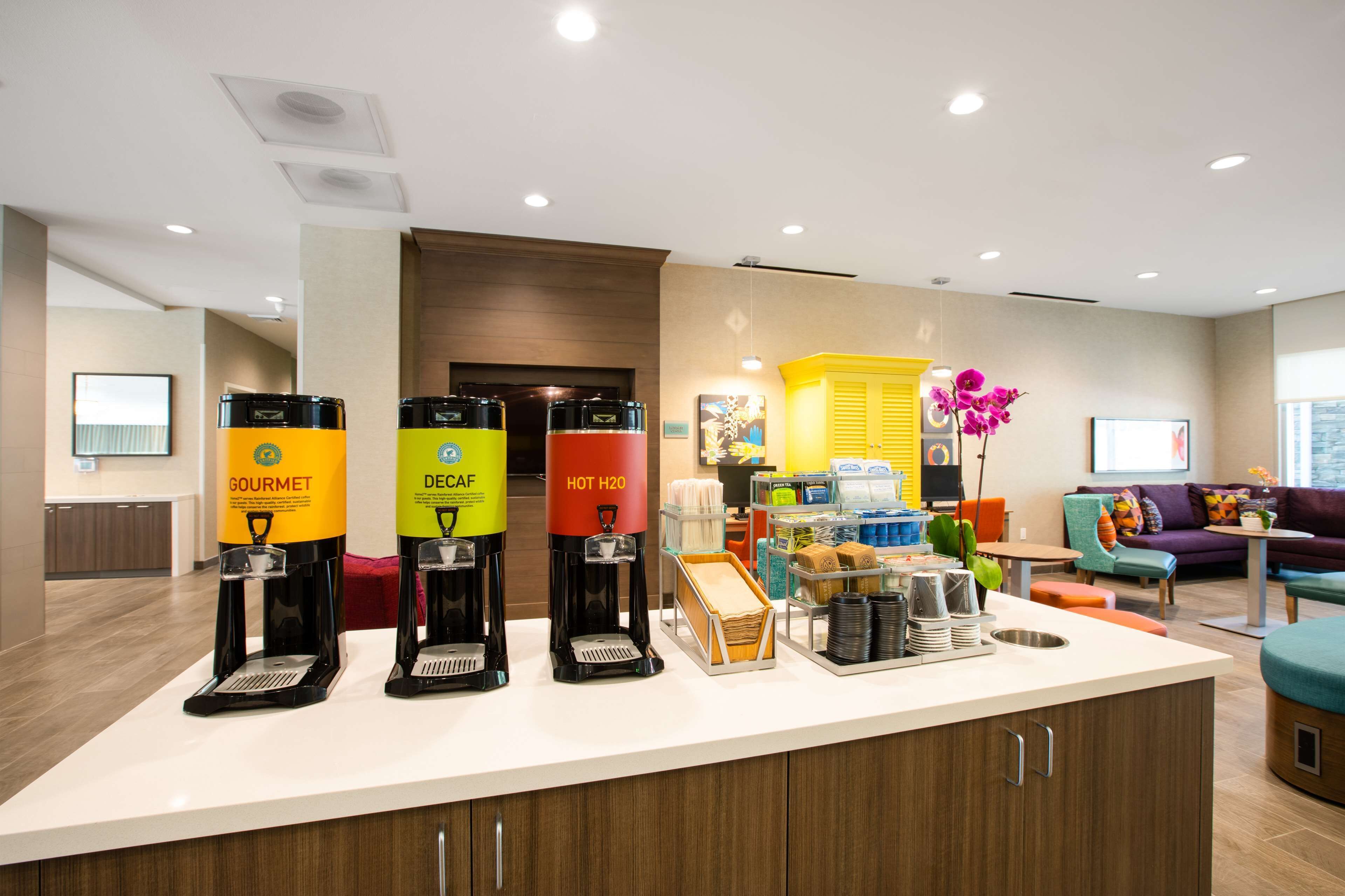 A modern hotel lobby with a coffee station featuring Gourmet, Decaf, and Hot H2O options.