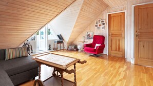 Et loftrum med skrå vægge, trægulv, sofa, rød lænestol, bord og en dør.