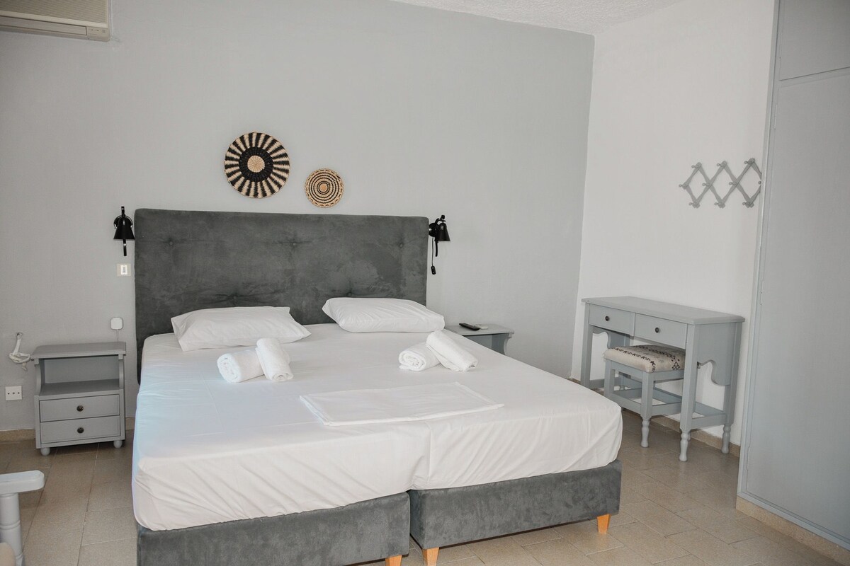 Una cama bien hecha con sábanas y almohadas blancas, un cabecero tapizado en gris y dos mesitas de noche con lámparas.