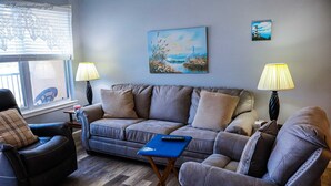 Una sala de estar con un sofá gris, dos sillones, una mesa de centro y dos lámparas. Hay un cuadro en la pared y una ventana con persianas.