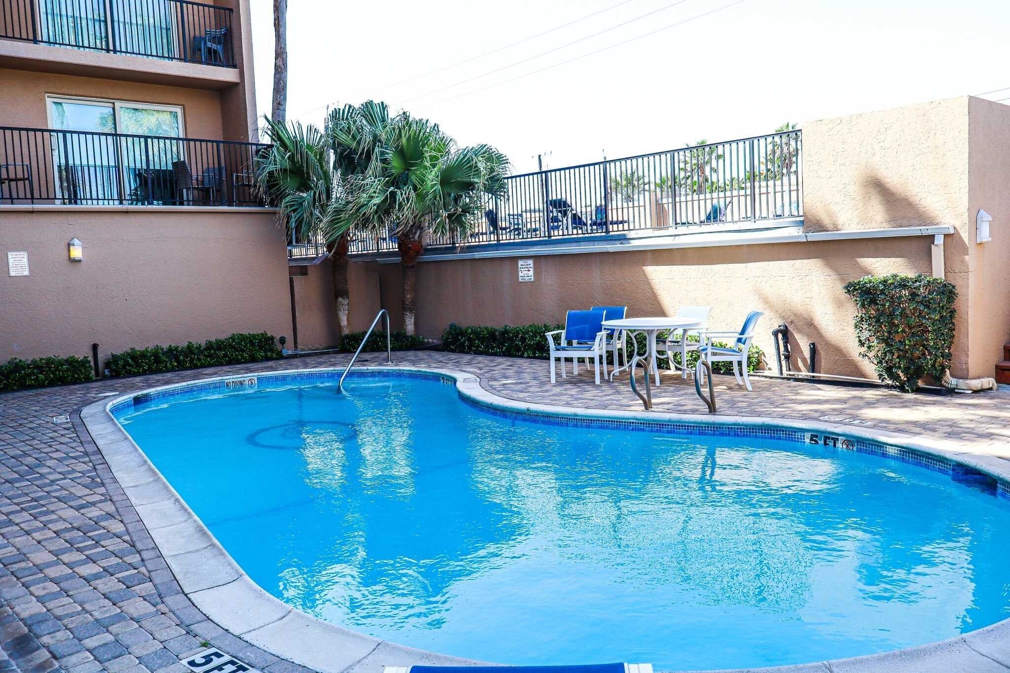 A swimming pool with a tiled deck, surrounded by lounge chairs and a small table.