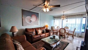 A living room with a brown leather sofa, a glass coffee table, a ceiling fan, a dining area with a table and chairs, and a large window with curtains.