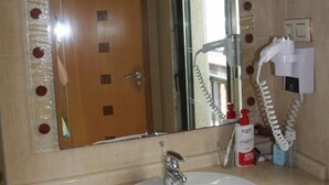 Une salle de bain avec un lavabo blanc, un miroir dont le reflet se superpose à une porte en bois, et un sèche-cheveux fixé au mur.