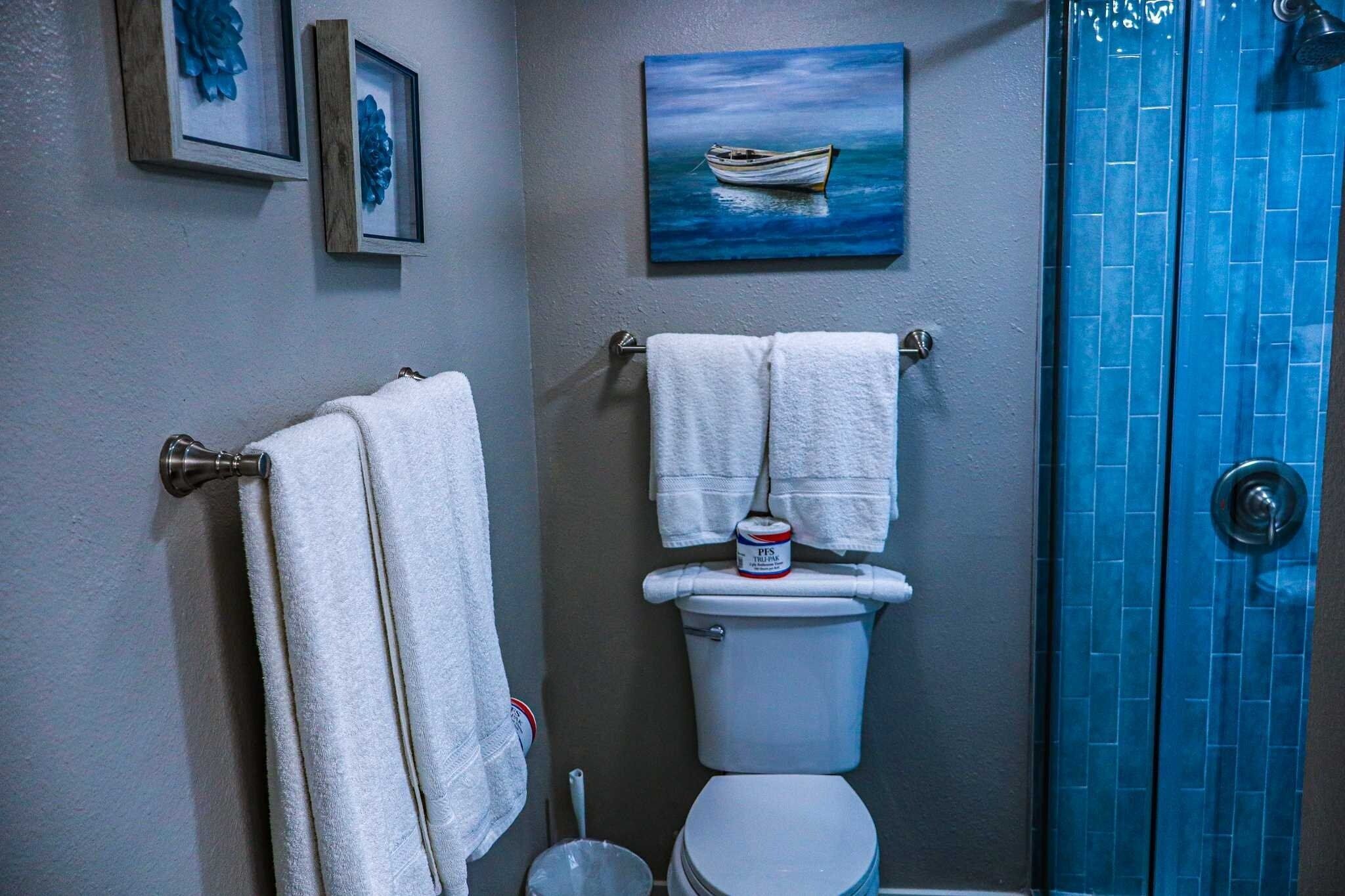A bathroom with a toilet, a blue shower curtain, and a painting of a boat on the wall.