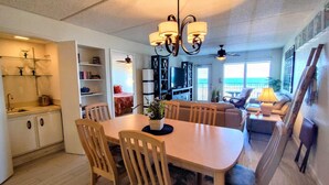 A spacious living area with a dining table, chairs, a sofa, and a TV. There is a kitchen area with cabinets and a sink. The room has a ceiling fan and a view of the ocean through large windows.