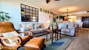 A spacious living room with a sofa, armchairs, and a coffee table. There is a kitchen area in the background.