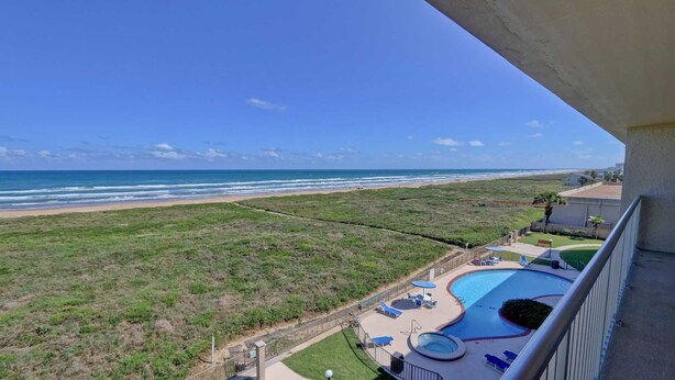 A balcony with a view of a beach and a swimming pool with lounge chairs. - Edgewater 507 Minium 3 Bedroom Condo by RedAwning (South Padre Island)