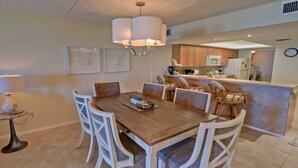 A dining area with a wooden table and white chairs, a chandelier, and a kitchen in the background. - Edgewater 507 Minium 3 Bedroom Condo by RedAwning (South Padre Island)