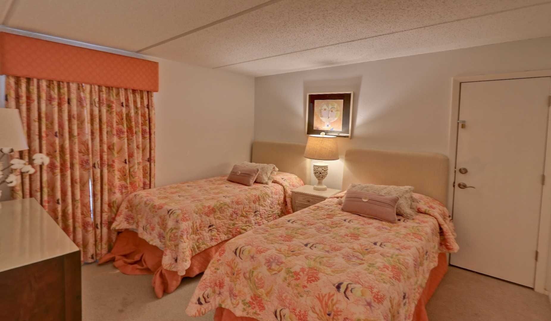 Habitación de hotel con dos camas, una mesita de noche con lámpara, una puerta y una ventana con cortinas.