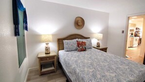 Un dormitorio con una cama, dos mesitas de noche, un sombrero colgado en la pared y una puerta que da a una cocina.