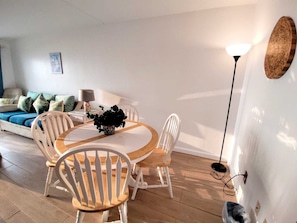 Ein modernes Wohnzimmer mit einem runden Holztisch, weißen Stühlen, einer Couch mit gemusterten Kissen, einer Stehlampe und einer Wanddekoration.