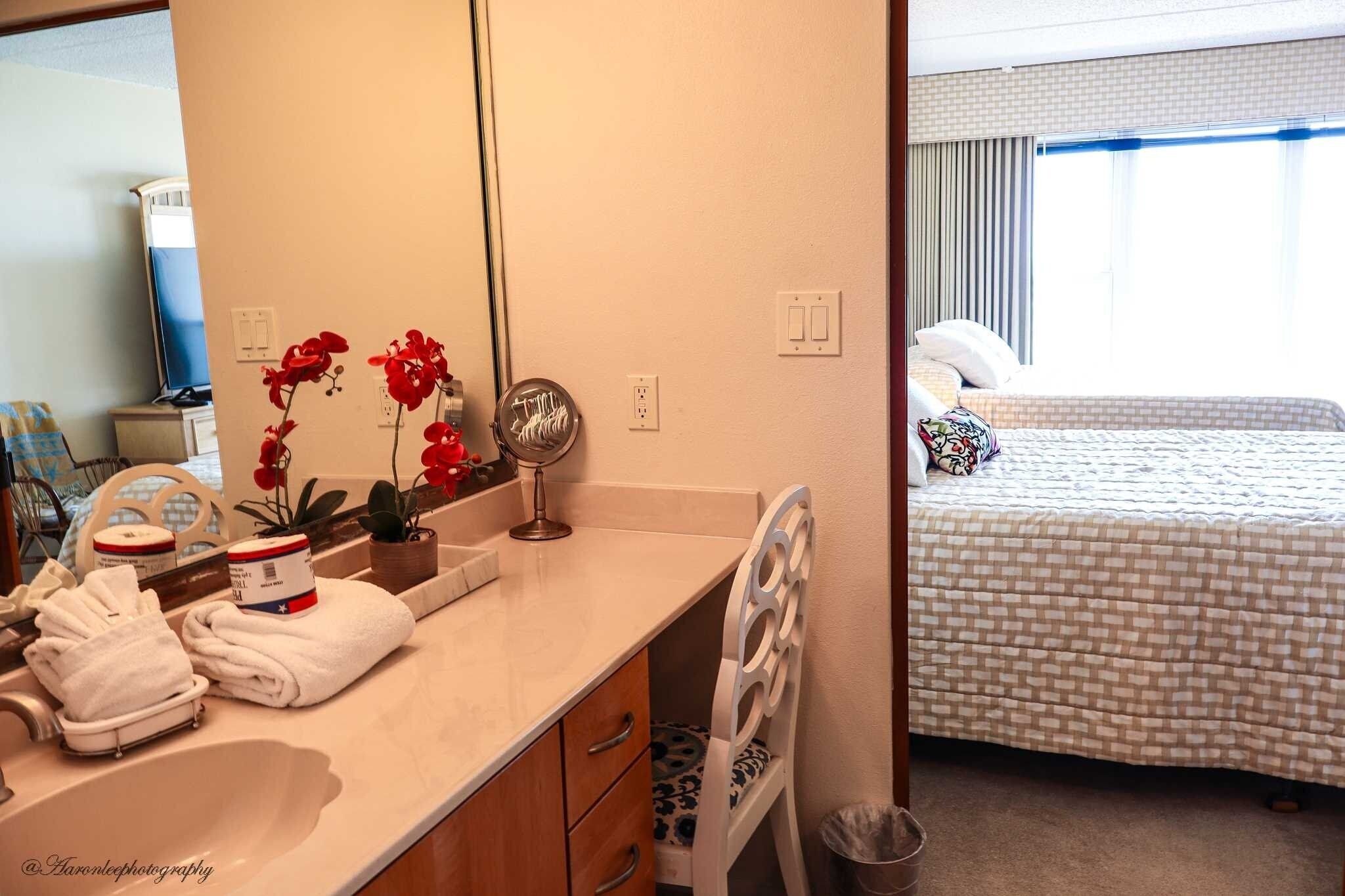 Une salle de bain d’hôtel avec un lavabo, un miroir et des orchidées rouges. La chambre adjacente contient un lit avec une couvre-lit à motifs et des oreillers.
