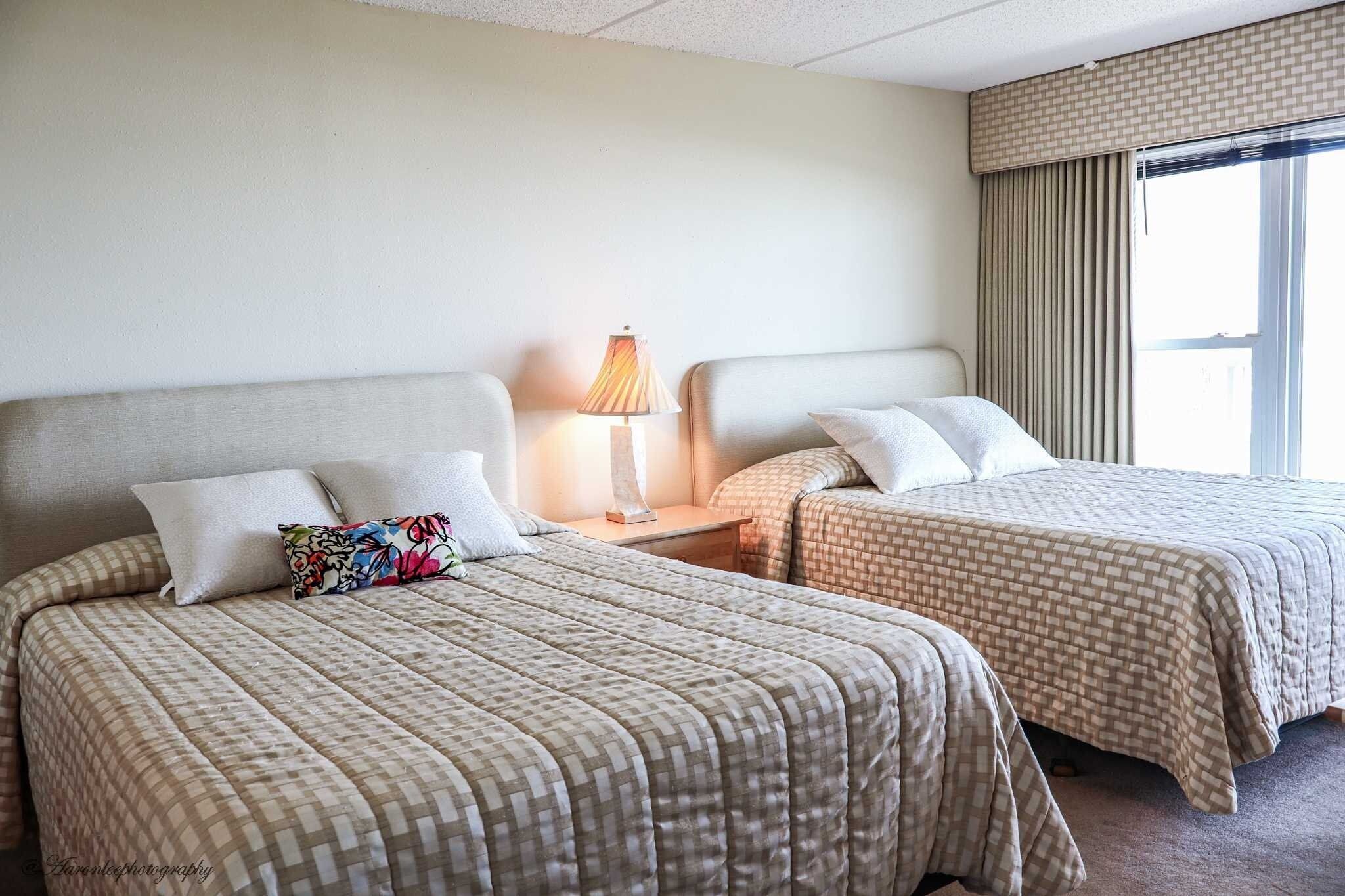 Una habitación de hotel con dos camas, cada una con una colcha a cuadros, una lámpara de noche y una ventana con cortinas.