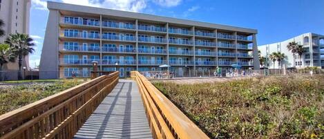 A multi-story hotel with balconies, a wooden boardwalk leading to the entrance, and a clear blue sky.