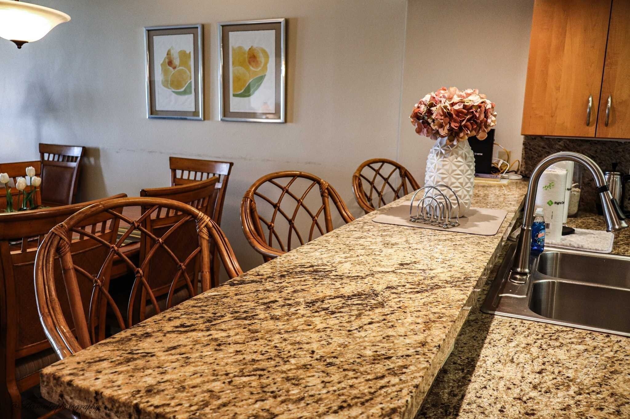 A kitchen with granite countertops, wicker chairs, a stainless steel sink, and framed artwork on the wall.
