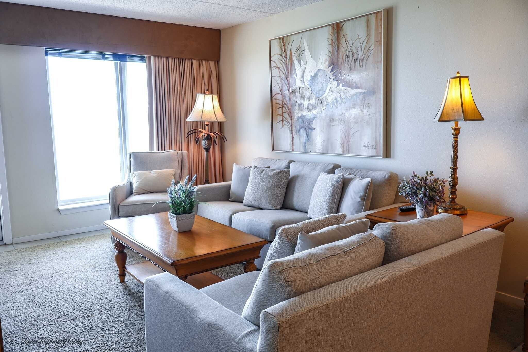 A living room with a sofa, armchair, and coffee table. There is a painting on the wall and a lamp on a side table.