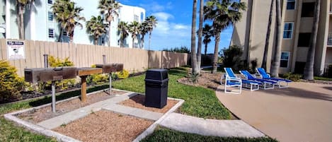 Zona de barbacoa con carbón designada, con un contenedor de basura, rodeada de palmeras y sillones de descanso.