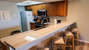 Une cuisine avec des armoires en bois, un réfrigérateur en acier inoxydable, un four à micro-ondes, un évier et un comptoir en marbre. Il y a trois tabourets au comptoir.
