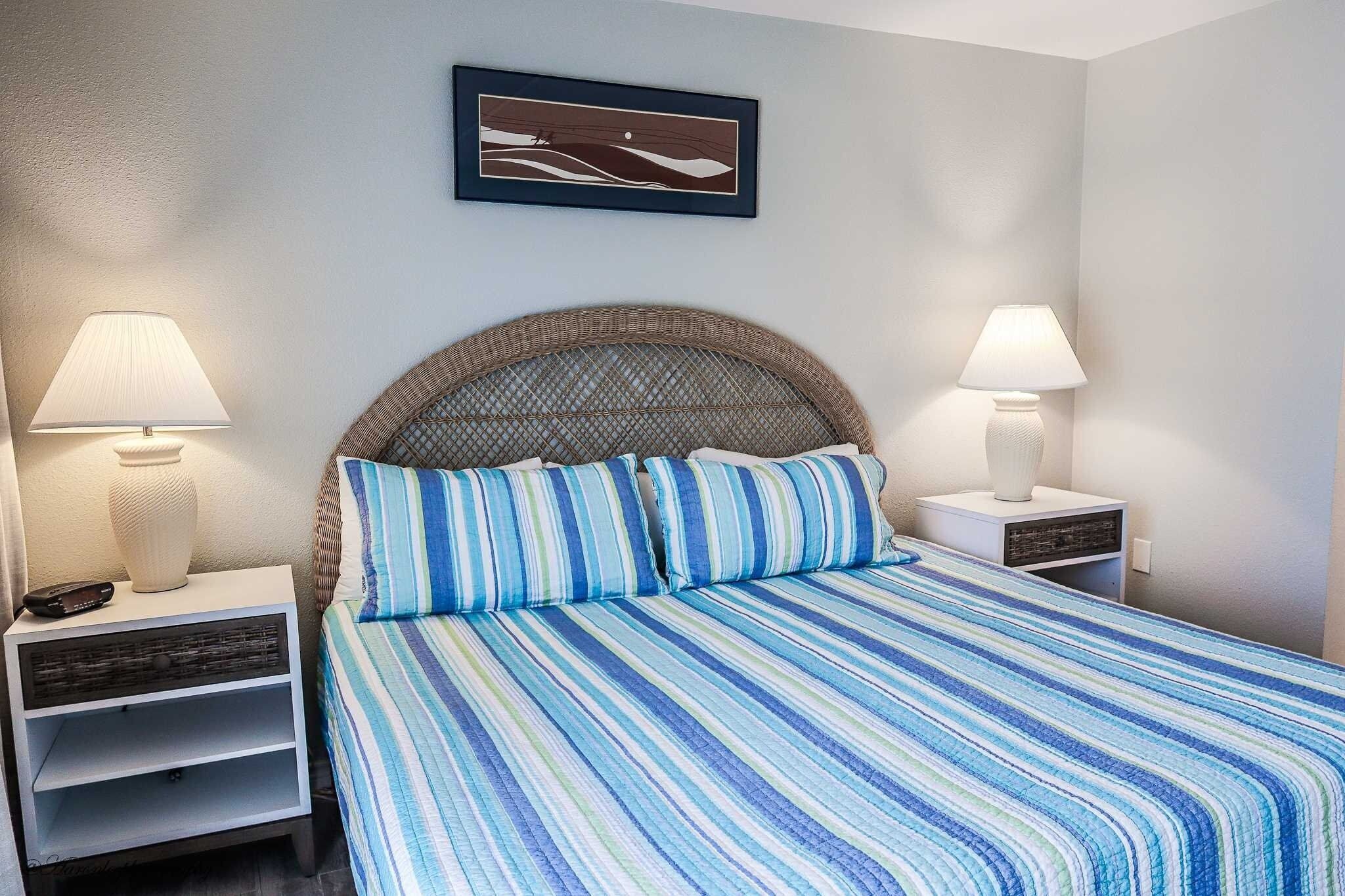A neatly made bed with striped bedding, two bedside lamps, a framed artwork on the wall, and a wicker headboard.