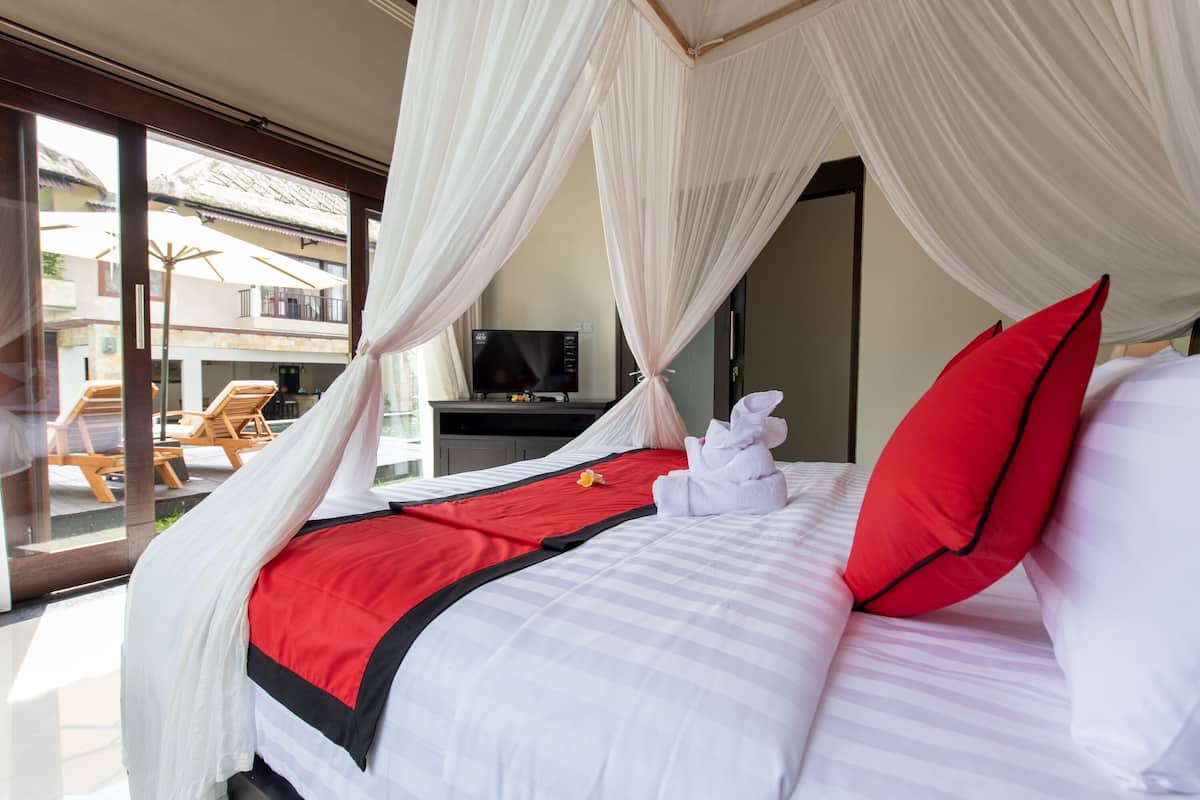 Un dormitorio con cama, cortinas blancas, colcha roja y negra, almohada roja y un televisor sobre un soporte.