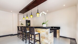 A modern kitchen with a stone island, dark wooden chairs, hanging pendant lights, and white orchids.