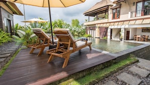 A wooden deck with lounge chairs and a pool, surrounded by tropical plants and a thatched-roof building.