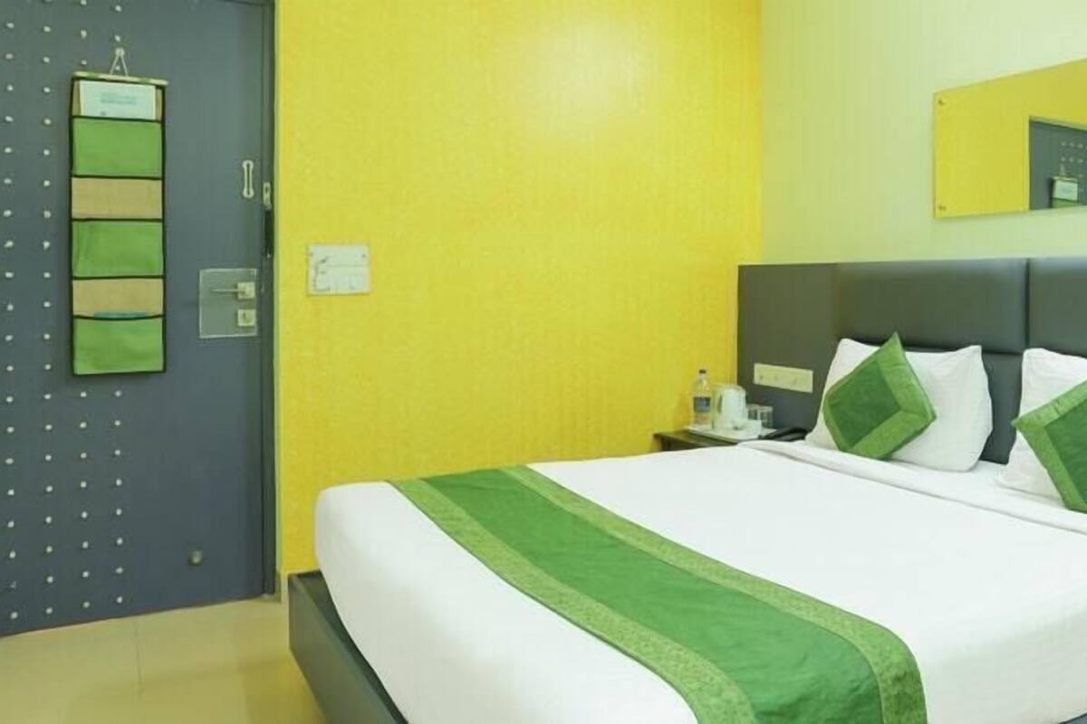Habitación de hotel con cama, mesita de noche, escritorio, silla, espejo y puerta con un tablón de anuncios.