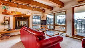 A cozy living room with a red leather sofa, a fireplace, and large windows offering a view of the mountains.