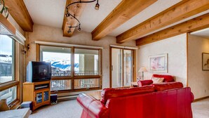 A living room with a red leather sofa, wooden beams, a television on a wooden stand, and a large window with a view of snow-capped mountains.