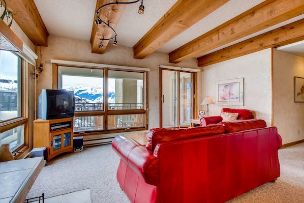 A living room with a red leather sofa, wooden beams, a television on a wooden cabinet, and a large window with a view of mountains.