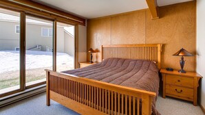 Een slaapkamer met een houten bed, twee nachtkastjes met lampen, een groot raam en uitzicht op een huis en tuin.