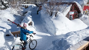 Een persoon op een fiets, met ski’s vastgemaakt aan de achterkant, rijdt over een besneeuwd pad met een houten hek en een rode gebouw op de achtergrond.