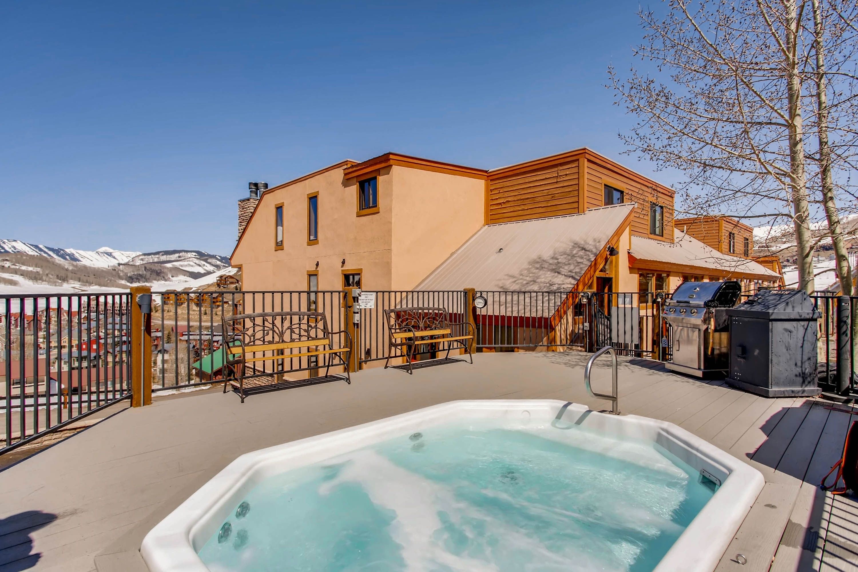 A hot tub on a deck with a view of mountains, a wooden building, and a fenced area with outdoor furniture.