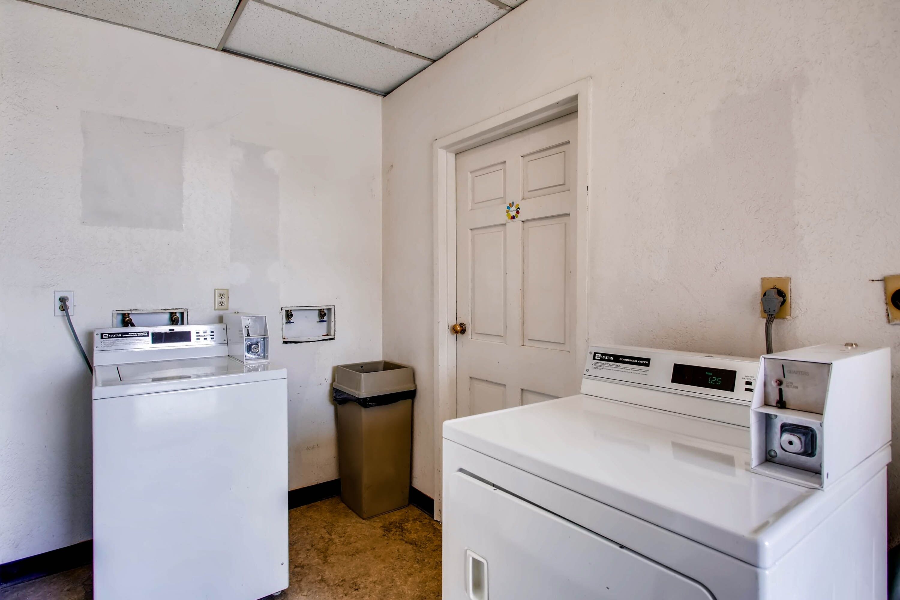 A laundry room with two white washing machines, a trash bin, and a door.
