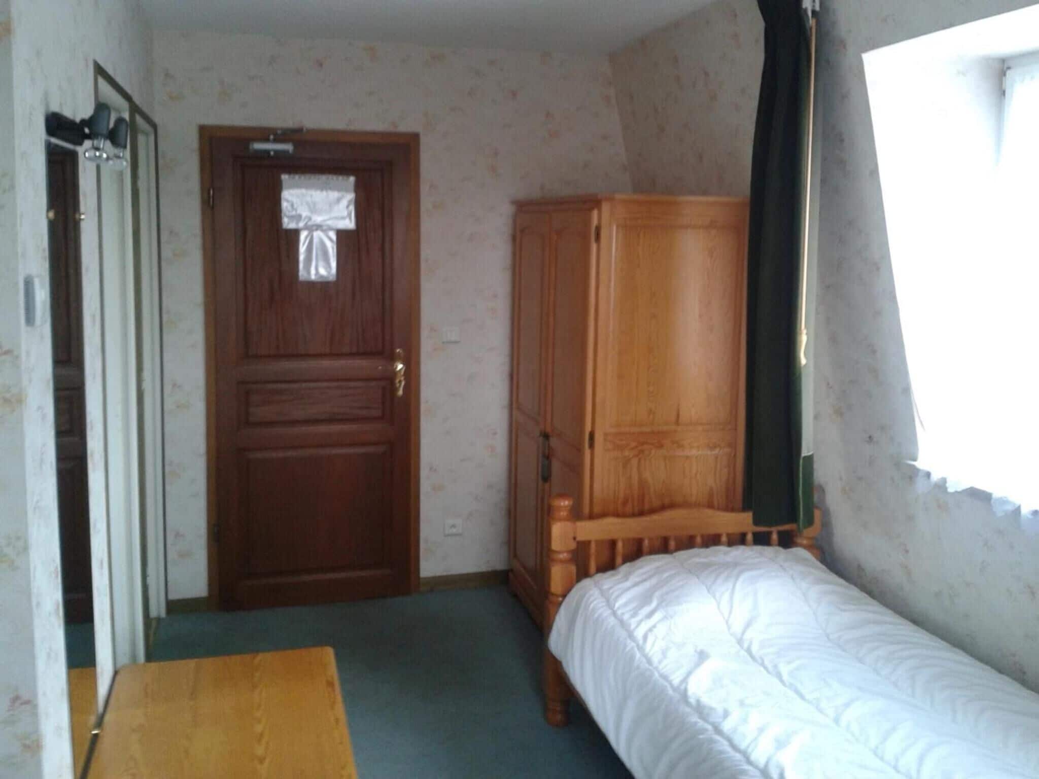 A small, simple room with a wooden door, a wooden wardrobe, a single bed, and a small table.