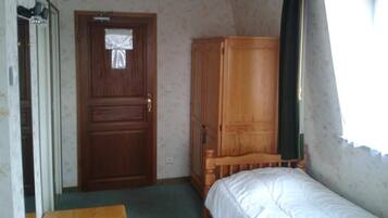 A small, simple room with a wooden door, a wooden wardrobe, a single bed, and a small table.
